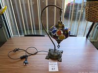 Complete view of the lamp on a wooden surface showing the curved metal arm, ornate base, colorful mosaic glass shade, and electrical cord with European plug.