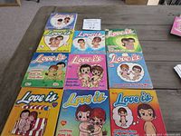 Top down view of 10 'Love Is' cartoon books numbered 2,4-12 laid out on a wooden table showing illustrated colorful covers with cartoon couple characters