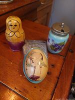 Photo showing wooden nesting doll, small violet pot with metal lid, engraved metal pot, painted decorative egg and its blue base on wooden surface.
