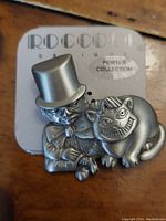 Front view of pewter brooch showing Mad Hatter with top hat and the Cheshire Cat side by side, mounted on Roccoco branded card.