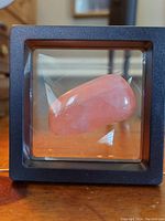 Front view of the rose quartz stone inside a black square glass frame on a wooden surface, showing the pink polished stone and clear glass sides.