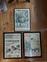 Top-down photo of three framed vintage newspaper covers laid flat on a wood floor. Each features headlines and photos about The Rolling Stones and other musical artists from the early 1960s.
