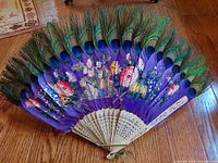 Front angle showing open fan with peacock feathers and detailed painted artwork on purple fabric, wooden base with carvings and painted details visible
