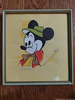 Frontal photo of the watercolor painting showing Mickey Mouse wearing a green hat and red scarf, framed in a light green frame.