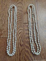 Two vintage cultured pearl necklaces laid out flat on a wooden floor. Each necklace is a single long strand of round pearls.