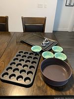 Baking items displayed on a wooden table including a mini muffin tin, round cake pans, small green bowls, cooling racks, cookie sheets, and heart-shaped cutters.