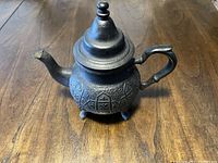 Front angled view of the dark cast iron tea kettle with ornate embossed design, spout, handle, and three legs on a wooden surface.