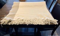 Cream-colored blanket neatly folded on a dark wooden table, showing fringed edges and soft texture.