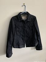 Dark blue denim jacket hanging on a white wall. Showing front button closure, collar, chest pockets with flaps and buttoned cuffs.