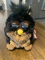 Front view of the black and brown 1998 Furby with big eyes, yellow beak, and intact fur sitting on a wood surface.