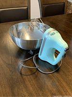 Light blue KitchenAid hand blender with metal beaters next to a stainless steel mixing bowl on a wooden table.