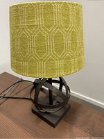 Photo of a table lamp with metal spherical base and green geometric fabric shade placed on a wooden surface against a white wall.
