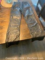 Front view of black leather pants laid flat on a wooden floor, showing full length and details such as waistband and zip fly.