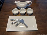 White ceramic rectangular serving dish displaying three stainless steel cheese spreaders and three white ceramic ramekins, all on a wood surface.
