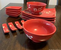 Red sushi dinnerware set arranged showing bowls, plates, dipping dishes, and chopstick rests with white flower patterns on a dark wooden table.