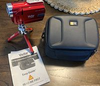 Red Vivitar DVR508NHD digital video camera on red mini tripod, accompanied by black carrying case and user manual on wooden table.