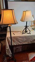 Photo of both tall floor lamp and shorter table lamp side by side on a covered table against a light background showing detail of bases and shades.