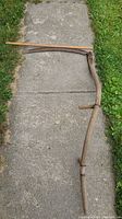 Full view of the antique scythe laid on concrete showing wooden handle, horizontal grip, leather binding and metal blade.