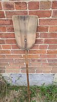 Full view of the all wood grain shovel leaning against a brick wall, showing the long handle and the wide wooden blade.