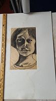 Frontal charcoal portrait on aged backing paper, showing a woman with short hair and prominent facial shadows. Ruler for scale included.