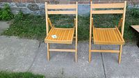 Two mid-century vintage folding maple chairs placed outdoors on a sidewalk in front of a stone wall. Chairs have slatted seats, a simple double horizontal slat backrest, and foldable design.