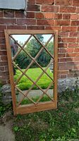 Full view of the wooden framed mirrored window with diamond shaped muntins, leaning against brick wall outside.