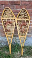 Full view of pair of Gros Louis snowshoes with wooden frames and leather straps leaning against brick wall.