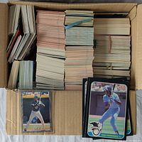 Top-down photo of open box filled with large stacks of baseball cards mostly Donruss from early 1980s with a few visible individual cards in protective sleeves.
