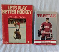 Both books displayed side by side showing front cover: 'Let's Play Better Hockey' by Ken Dryden with red cover and photo of Dryden in goalie gear, and 'Tretiak the Legend' by Vladislav Tretiak with dust jacket showing Tretiak in Soviet CCCP jersey.