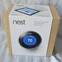 The unopened original cardboard box of the Nest Learning Thermostat showing product branding and image of thermostat display on sleeve.