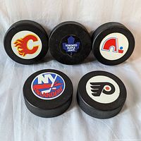 Five NHL hockey pucks with team logos of Calgary Flames, Toronto Maple Leafs, Quebec Nordiques, New York Islanders, and Philadelphia Flyers.
