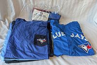 Image of various folded blue Toronto Blue Jays T-shirts and one replica jersey with a visible Blue Jays logo patch.