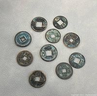 Top-down view of 10 old Chinese metal Feng Shui coins arranged in a loose pattern on light leather surface. Coins are round with square holes through the center, showing some green patina and rust. Various Chinese characters visible on each coin.