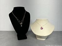 Three necklaces on display busts including a multi-layer necklace on black bust and a silver necklace with purple heart on white bust