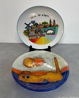 Two decorative plates: foreground is a fused glass plate with abstract warm tones, background is a ceramic plate with painted colorful Jerusalem cityscape and Hebrew text.