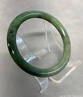 Natural green jade bangle shown on clear plastic stand from angle highlighting polished surface and color variations.