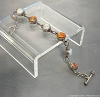 Full view of the bracelet laid on a transparent display stand showing alternating white and orange/brown pearls with silver links.