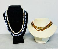 Side by side display of four necklaces: Chinese blue and white porcelain, braided bead necklace, tiger's eye, amber bead necklace.