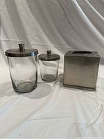 Front view of glass containers with brushed nickel lids and square tissue box cover in brushed nickel finish.