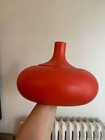 Red rounded pendant light shade with neck narrowing upwards, held in hand against plain wall background.