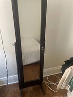 Front view of closed jewelry storage cabinet with full length mirror on door showing the cabinet standing on wooden floor against a white wall.