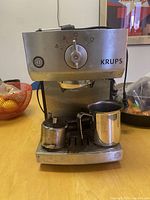 Photo of Krups espresso machine on table with small milk frothing pitcher and coffee container in front.