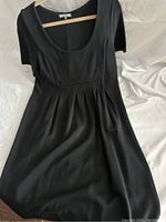 Black short-sleeve dress hanging on wooden hanger, showing full front view with pleats and scoop neckline
