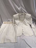 White cotton suit consisting of fitted jacket with long sleeves and matching skirt with beige belt, displayed on hangers