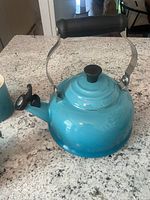 Close-up front view of teal Le Creuset tea kettle with black handle and spout cap on granite countertop.