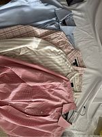 Four men's dress shirts laid out showing colors and patterns including light blue, beige plaid, cream stripes, and pink stripes.