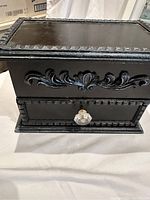 Black wooden jewellery box showing ornate carved design and glass knob drawer from front.