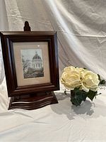 Dark brown wooden picture frame with white mat holding a 5x7 black and white image of a building dome, next to a small vase with cream silk flowers.