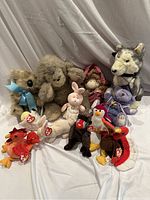 Photo showing assorted stuffed toys including Beanie Babies such as a monkey, wolf, bunny, doll, and various Beanie Babies: lobster, elephant, goat, cardinal bird, and penguin.