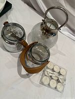 Two glass candle holders, one with a metal handle, one with a leather strap, and a blister pack of tealight candles on a cloth surface